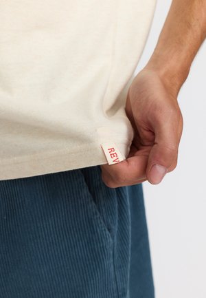 Beige katoenen t-shirt met een rood label met de tekst "REV" aan de onderrand, vastgehouden door een hand. De achtergrond is eenvoudig en licht.