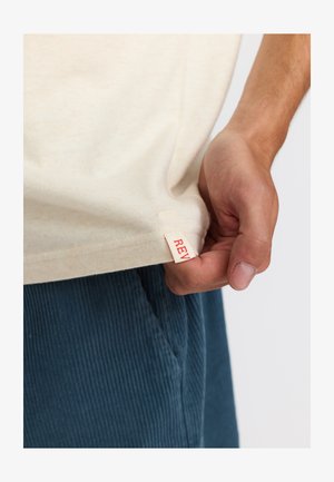 Beige katoenen t-shirt met een rood label met de tekst "REV" aan de onderrand, vastgehouden door een hand. De achtergrond is eenvoudig en licht.