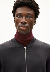 Schwarzer Reißverschluss-Pullover aus strukturiertem Stoff, darunter ein eng anliegender weinroter Rollkragen und metallische Reißverschlussdetails.