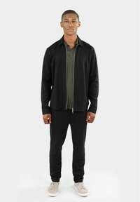 Schwarze Zip-Jacke über einem grünen Poloshirt, kombiniert mit schwarzen Hosen und hellgrauen Sneakers. Glatter Stoff, taillierter Schnitt, minimalistischer Stil.