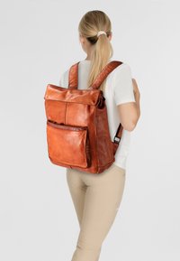 Zaino in pelle marrone con pattina superiore, tasca frontale e spallacci regolabili. Texture liscia e design minimalista.