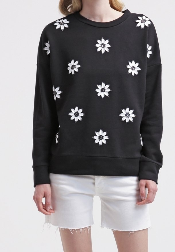 Personne portant un haut noir à manches longues avec des motifs de marguerites blanches, associé à un short en jean blanc effiloché.