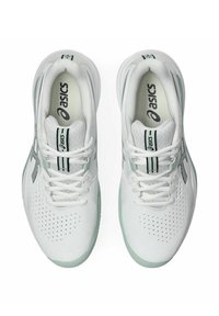 Vita atletiska skor med en slät läderöverdel, perforerade detaljer, gröna accenter på sulan och svart logotyp längs sidorna.