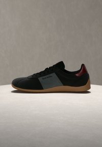 Basket basse noire avec semelle en gomme, panneau latéral gris et accent talon rouge foncé, exposée sur une surface claire avec un fond neutre.