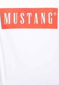 Camisa blanca de algodón con un gran gráfico rectangular rojo que lleva la palabra "MUSTANG" en letras blancas y negrita.