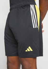 Pantalones cortos deportivos negros con cinturilla elástica, que presentan detalles en amarillo y tres franjas verticales blancas en el lateral. Diseño de tejido texturizado.