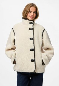 Chaqueta de forro polar color crema con cuello alto, cierres de botones negros y ribetes en contraste. Presenta un ajuste extragrande y bolsillos laterales.