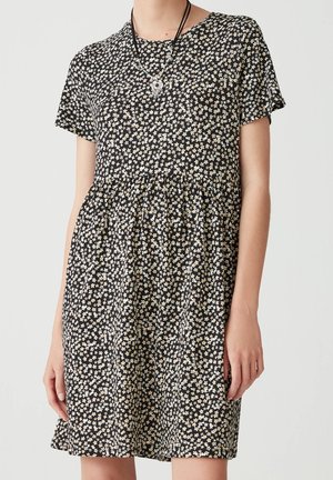 Woman wearing a short-sleeve black dress with a small white and beige floral pattern and layered silver and black necklaces.