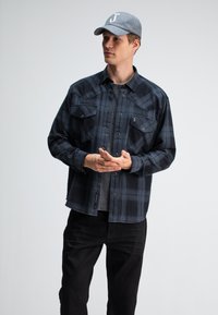 Zwarte en marineblauwe geruite overhemd met lange mouwen en twee borstzakken, drukknoopsluiting, gedragen over een grijs T-shirt, gecombineerd met zwarte broek en een grijze pet.