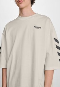 Korte mouwen, beige katoenen t-shirt met een zwart "hummel sportswear" logo en hoekige zwarte strepen op de mouwen. Gladde textuur.