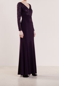 Lange paarse jurk met een V-hals, voorzien van een pailletten lijfje en lange mouwen. De soepele rok vloeit naar de grond met een getailleerd silhouet.