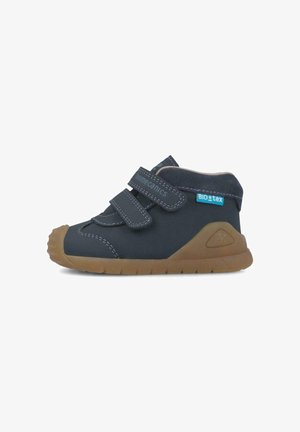 Scarpe per bambini di colore blu navy con due cinturini in velcro e cuciture grigie a contrasto. Presentano una suola in gomma marrone flessibile e un'applicazione testurizzata.