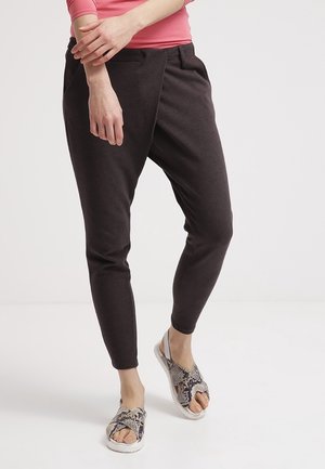 Kvinde iført mørkegrå smalle bukser, lyserød langærmet skjorte og åben tå-sandaler med slangeskindsmønster, stående mod en ensfarvet baggrund.