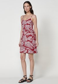 Rotes ärmelloses Kleid mit weißem Blumenmuster und Blattmotiven, aus leichtem Stoff gefertigt. Es hat ein tailliertes Design und einen ausgestellten Saum.