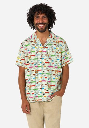 Homme aux cheveux bouclés souriant, portant une chemise à manches courtes avec un motif de carrés colorés du plateau de jeu Monopoly, et un pantalon beige.
