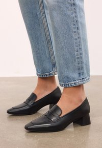Svarta läder loafers med blockklack och spetsig tå, prydda med en dekorativ rem över ovansidan. Bärs tillsammans med uppvikta ljusblå denimjeans.