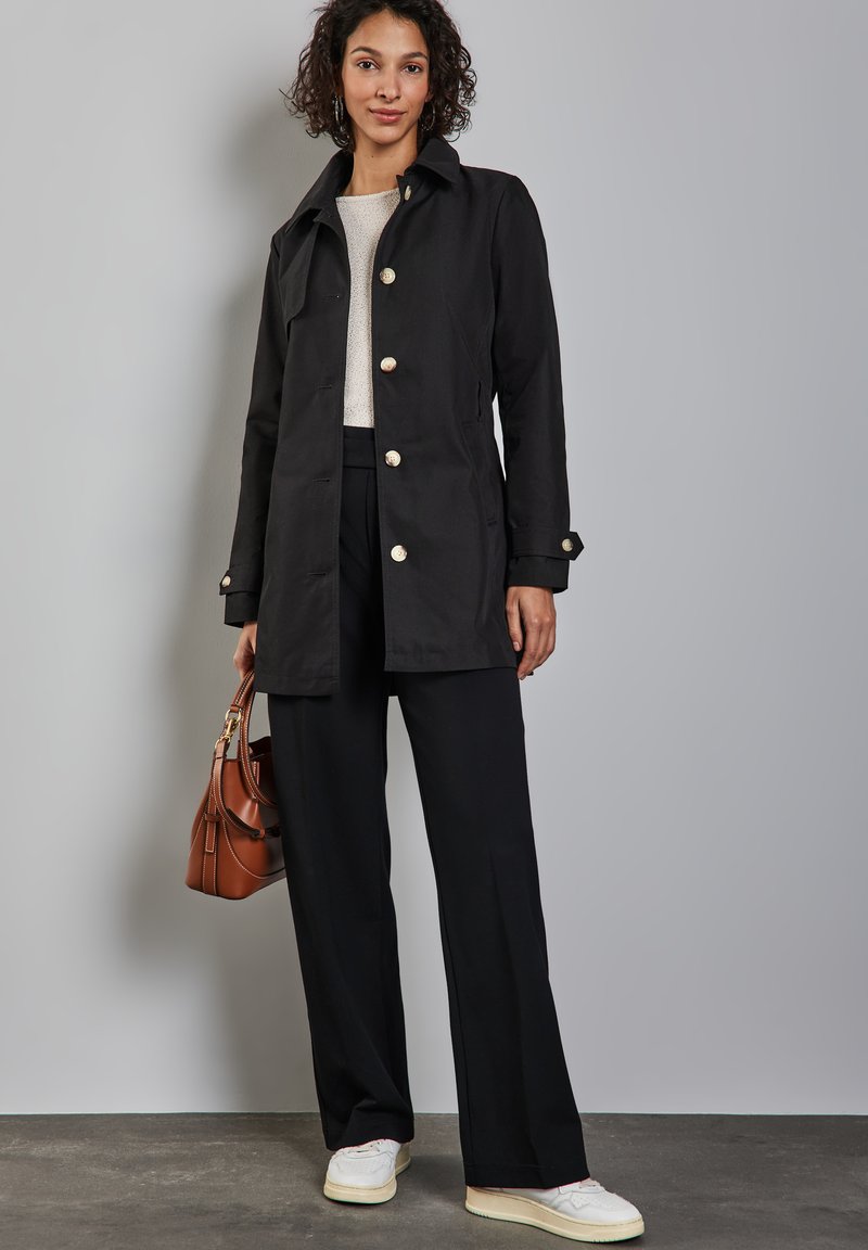 Czarny, długi płaszcz z długimi rękawami i dużymi guzikami, noszony na jasny sweter i dopasowane czarne spodnie. Dodatki to brązowa torba i białe sneakersy.