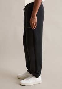 Schwarze Jogginghosen aus weichem Stoff mit einem elastischen Bund und Kordelzug, versehen mit einem Logodruck an der Seite. Kombiniert mit weißen Sneakern.