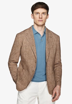 Homme portant un blazer marron texturé sur un polo bleu et un pantalon blanc, debout avec une main dans la poche sur un fond uni.