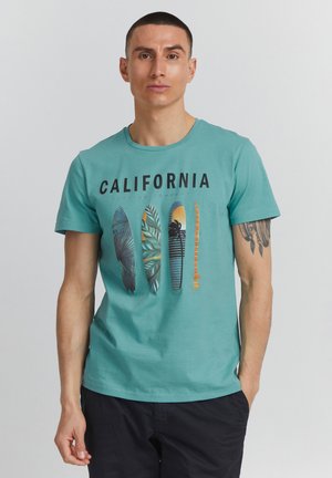 Rövid hajú férfi, aki türkizkék pólót visel, amelyen a "CALIFORNIA" felirat és négy szörfdeszka grafikája látható, fekete rövidnadrágban áll egyszínű háttér előtt, és karján tetoválás van.