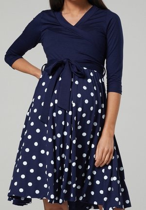 Femme portant une robe portefeuille bleu marine à manches trois-quarts avec une jupe à pois blancs, nouée à la taille avec un nœud.