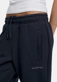 Marineblå joggebukser med elastisk liv og snøring. Har sidelommer og "CASPARA" -logo i lys grå broderi. Glatt stoffstruktur.