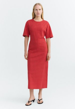 Frau steht, trägt ein rotes kariertes Midikleid mit kurzen Ärmeln und schwarze Sandalen mit Absatz, schlichter heller Hintergrund.