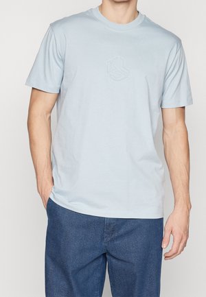 Camiseta de algodón azul claro con cuello redondo y mangas cortas, que presenta un logo hexagonal en relieve en el pecho. Corte casual con una textura suave.