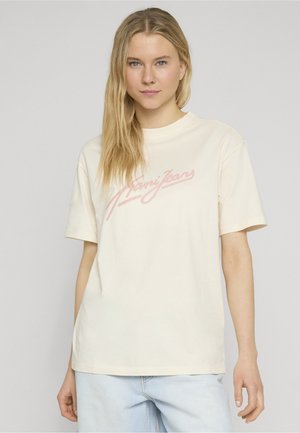 Blonde Frau mit blauen Augen, die ein cremefarbenes oversized T-Shirt mit pinker Schrift „Nani Jeans“ und hellblaue Jeans vor grauem Hintergrund trägt.