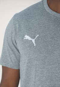 Grå kortermet t-skjorte laget av mykt stoff, med en hvit brodert Puma-logo på venstre bryst. Klassisk rund halslinning.