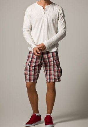 Chemise henley blanche à manches longues, bermuda cargo à carreaux rouge, bleu et blanc, et baskets rouges. Tenue décontractée avec une coupe ample.