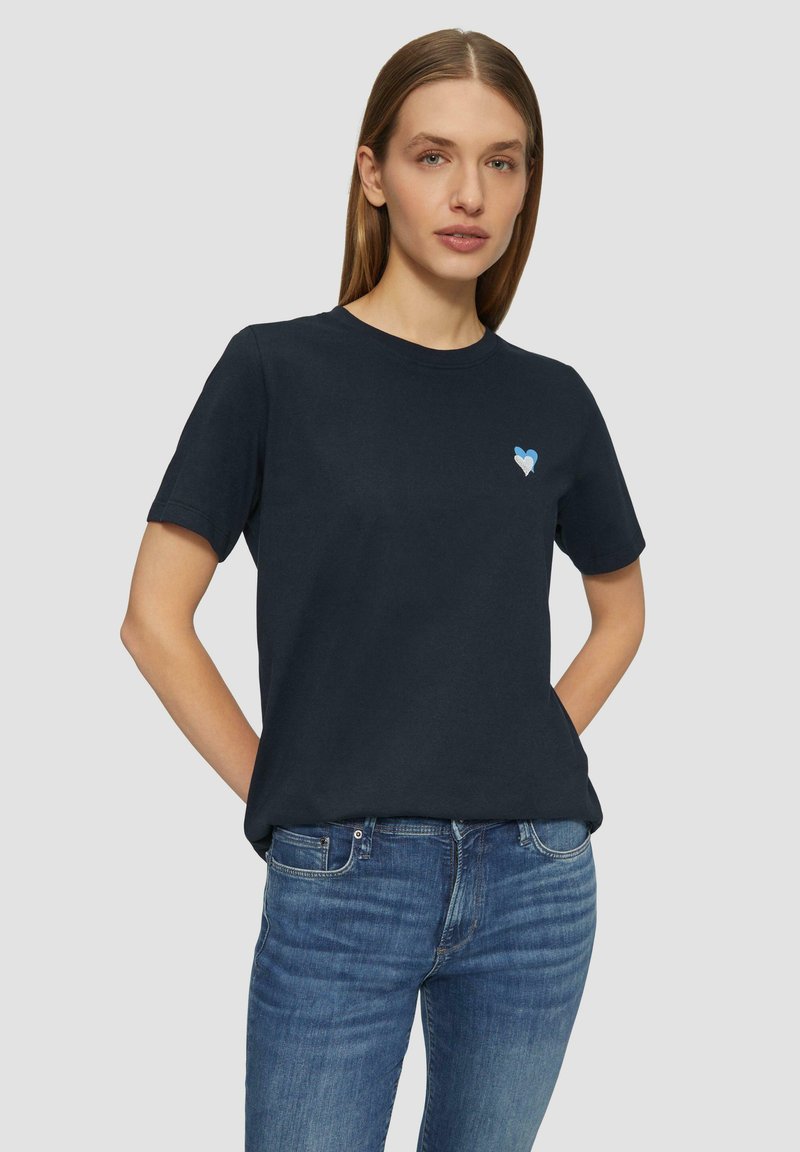 Marineblauwe katoenen t-shirt met een ronde halslijn, korte mouwen en een lichtblauw hartembleem op de linkerborst. Een paar blauwe spijkerbroeken.