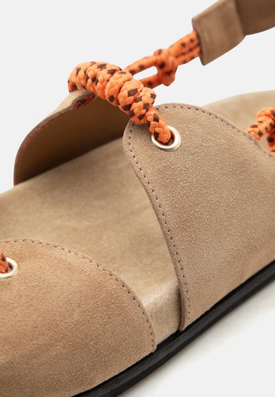 Μπεζ σουέντ σανδάλι με χονδρά πορτοκαλί και μαύρα πλεγμένα κορδόνια που περνούν μέσα από μεταλλικά ροδέλες σε μαύρη σόλα.
