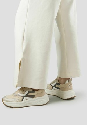 Primo piano di sneakers beige e bianche con suole spesse indossate con pantaloni crema a gamba larga con spacchi laterali.