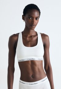 Weißer Sport-BH mit einem breiten elastischen Bund, der mit einem schwarzen "Calvin Klein"-Logo versehen ist. Das Design hat eine runde Ausschnittform und eine glatte Textur.