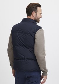 Marineblå dunvest med høy krage, kviltet design og polstret tekstur, båret over en beige langermet skjorte.