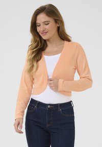 Kort, långärmad kofta i mjukt persikofärgat tyg, med öppen framkant och rundad sidsöm, tillsammans med en vit topp och mörka jeans.
