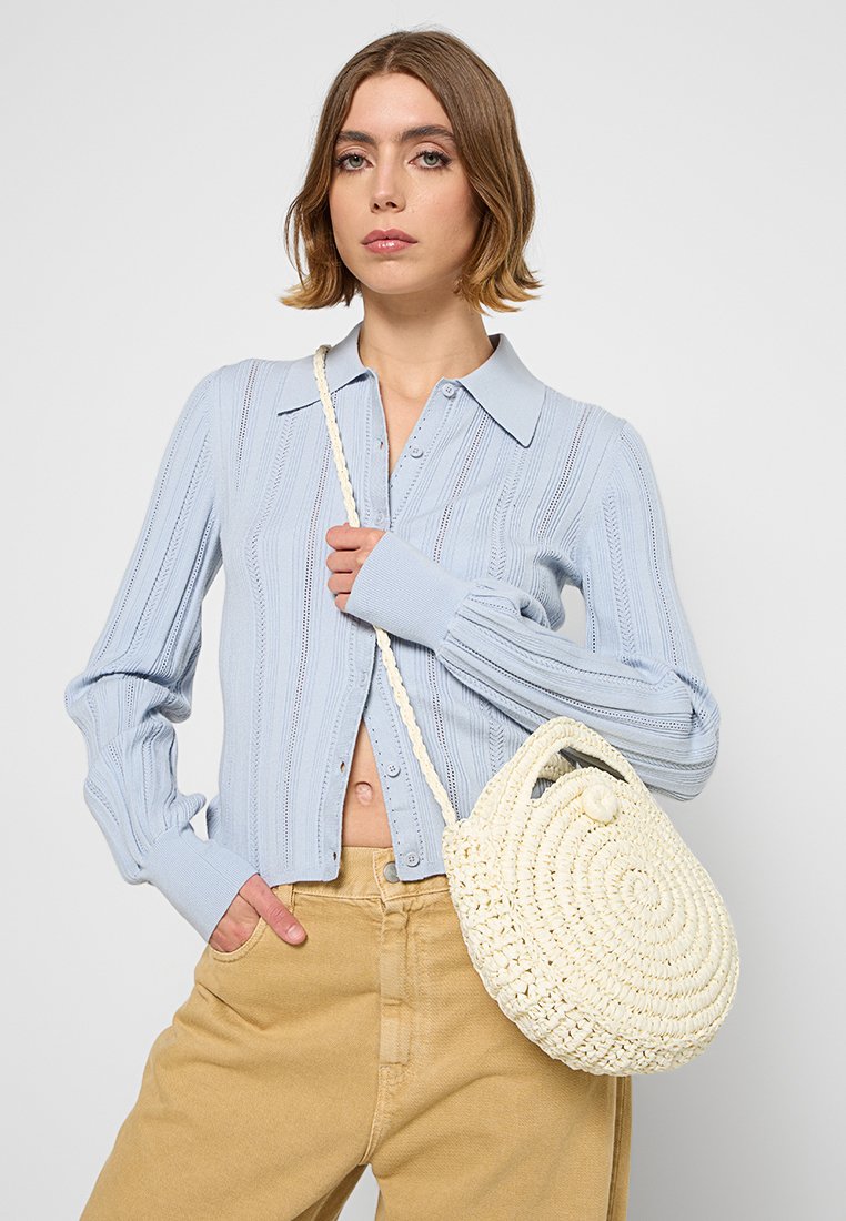 Cárdigan de punto azul claro con textura acanalada y cierre de botones, combinado con un bolso redondo de color crema tejido. Modelo vistiendo pantalones beige.