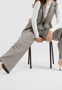 Šedý kostkovaný oblek se širokými kalhotami a střiženou vestou. Pod tím bílý žebrovaný dlouhý rukáv; outfit doplňují béžové podpatky.