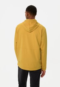 Gelbe Kapuzenjacke mit strukturiertem Finish, Taschendetails vorne und verstellbarer Kapuze. Kombiniert mit schwarzen Hosen für einen kontrastierenden Look.