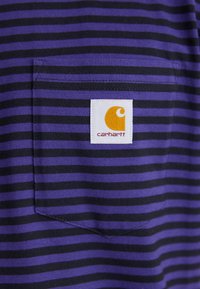 Violet un melna svītrainā T-krekls ar kabatu; tam ir balta birka ar oranžu Carhartt zīmolu. Kokvilnas materiāls, vienkāršs dizains.
