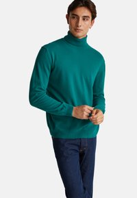 Turkusowy sweter z kołnierzem typu golf wykowany z miękkiej tkaniny, o dopasowanym kroju, z prążkowanymi mankietami i jednolitą strukturą, w zestawieniu z ciemnymi dżinsami.