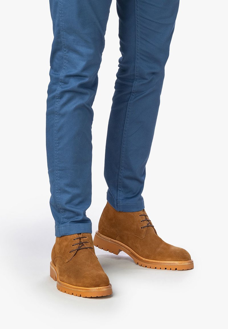 Stivaletti alla caviglia in suede marrone con design allacciato, suola in gomma e suola esterna in tono contrastante beige, abbinati a pantaloni aderenti blu.
