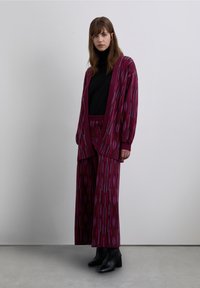 Magenta patterned knit cardigan over a black turtleneck, paired with wide-legged pants of the same design, complemented by black heeled boots.