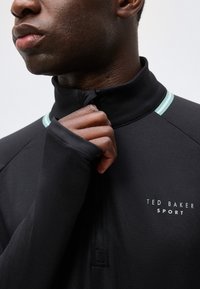 Κοντινή λήψη ενός ατόμου που κλείνει το φερμουάρ στον γιακά ενός μαύρου πουλόβερ Ted Baker Sport με γαλάζιο περιθώριο κοντά στους ώμους.