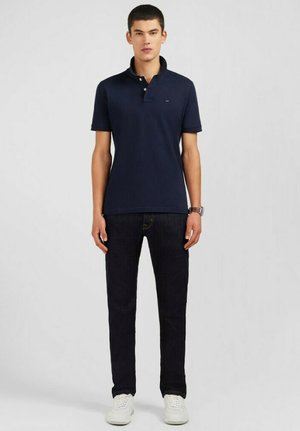T-shirt polo marine avec un col, des manches courtes et un logo subtil. Associé à un jean foncé et des baskets blanches, créant un look décontracté.