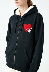 Zwarte zip-up hoodie met een pluizige witte voering, voorzien van een rode hart patch en veiligheidsspeld ontwerp op de linkerborst, kangoeroezakken.