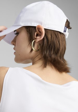 Cappello bianco in cotone con visiera curvata, dotato di una fascetta regolabile sul retro e di una piccola etichetta con logo. Orecchino a cerchio grande in oro visibile.