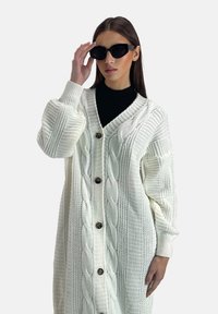 Weißer Strickcardigan mit Zopfmuster, lässiger Passform, V-Ausschnitt und Holzknöpfen. Getragen über einem schwarzen Rollkragenpullover, kombiniert mit schwarzen Sonnenbrillen.