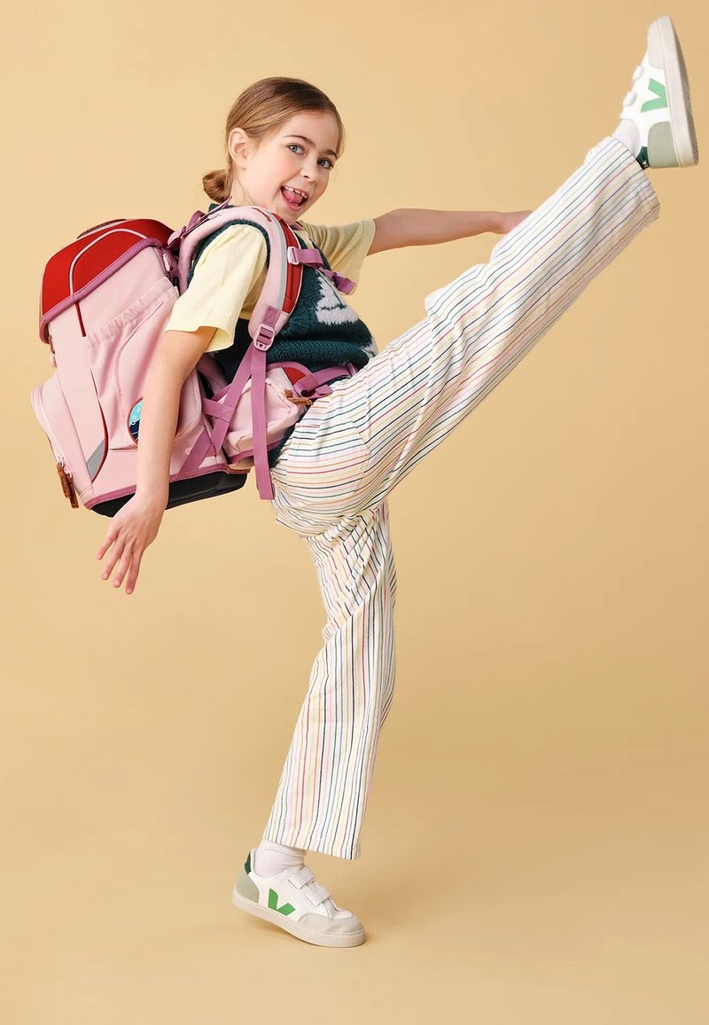 Zaino rosa e rosso con spallacci regolabili, indossato da un bambino. Pantaloni a righe multicolori e scarpe bianche con accenti verdi.